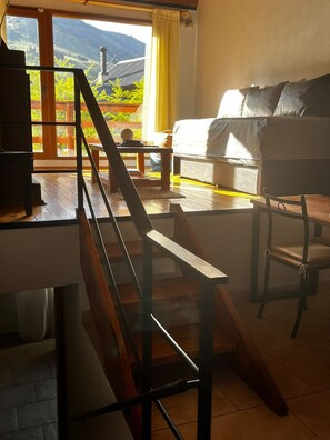 A room with a wooden staircase, a sofa, a table, and a view of a mountain landscape through large windows. - VF Catedral Mountain Department (San Carlos de Bariloche)