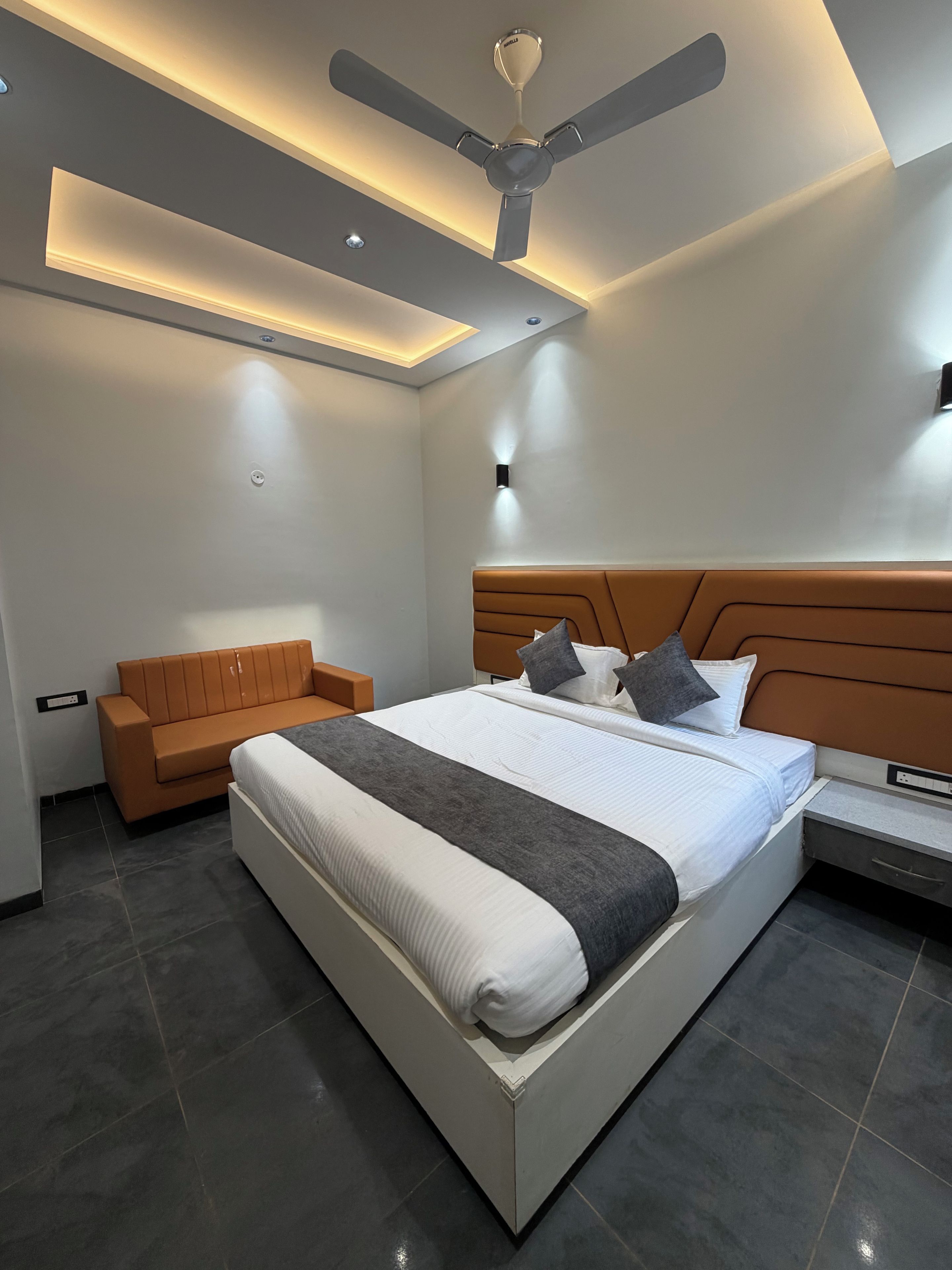 A modern bedroom with a large bed, a nightstand, a sofa, and a ceiling fan.