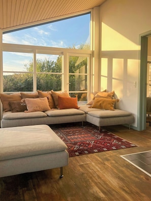 A modern living room with a sectional sofa, a patterned rug, and large windows offering a view of greenery. - Idyll and charm with views over the fjord (Kalundborg)
