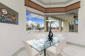 A dining area with a glass table and white chairs, a painting on the wall, and a view of palm trees and buildings outside. - Ember Noir | Waterfront Pool & Spa | South End (Marco Island)
