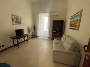 A living room with a sofa, a television on a stand, a bookshelf, and a window with curtains. - Vatican Doli (Roma)