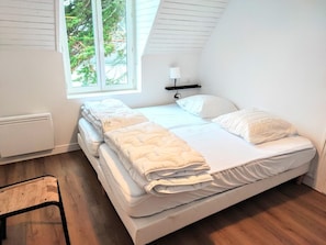 Una cama individual con colchón y almohadas blancas, una lámpara de noche y una ventana con vista a árboles.