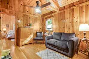 Een gezellig chaletinterieur met een houten plafond, een plafondventilator, een bank, een stoel, een klein tafeltje en een lamp.