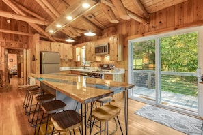 A wooden cabin interior with a central island bar, bar stools, a kitchen with wooden cabinets, and a sliding glass door leading to an outdoor deck. - Riverside Cabin, Mtn Views, Fire Pit and Fireplace (West Jefferson)