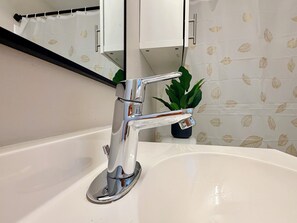 A bathroom with a chrome faucet, a white sink, a potted plant, and a mirror with a leaf-patterned wall. - Stylish Studio in St Boniface: French Quarter - cafés, museums, bus route, park (Winnipeg)
