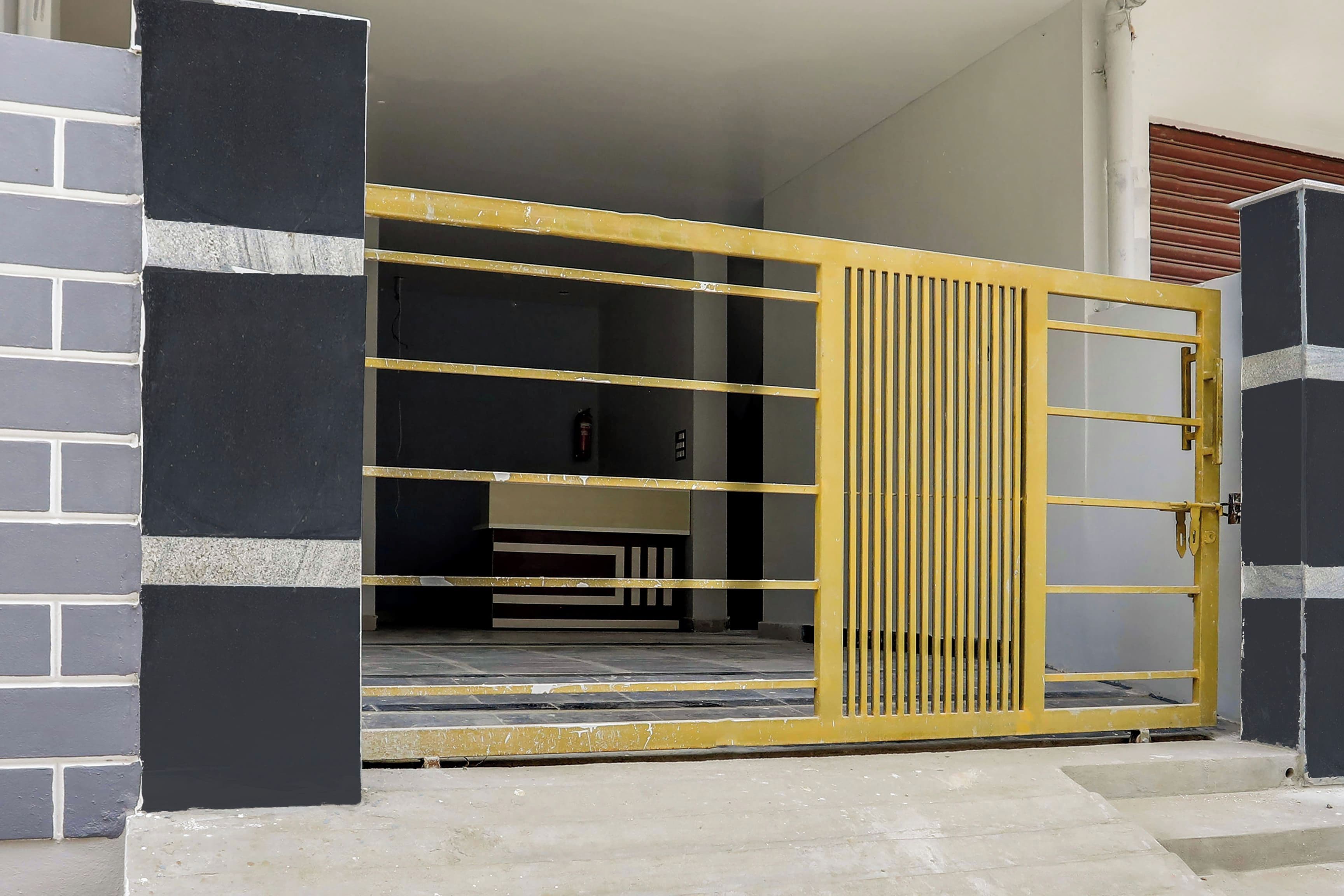 Una entrada protegida por un portón de metal amarillo, un camino de hormigón que lleva a la entrada de un edificio, y un muro de ladrillo a la izquierda.
