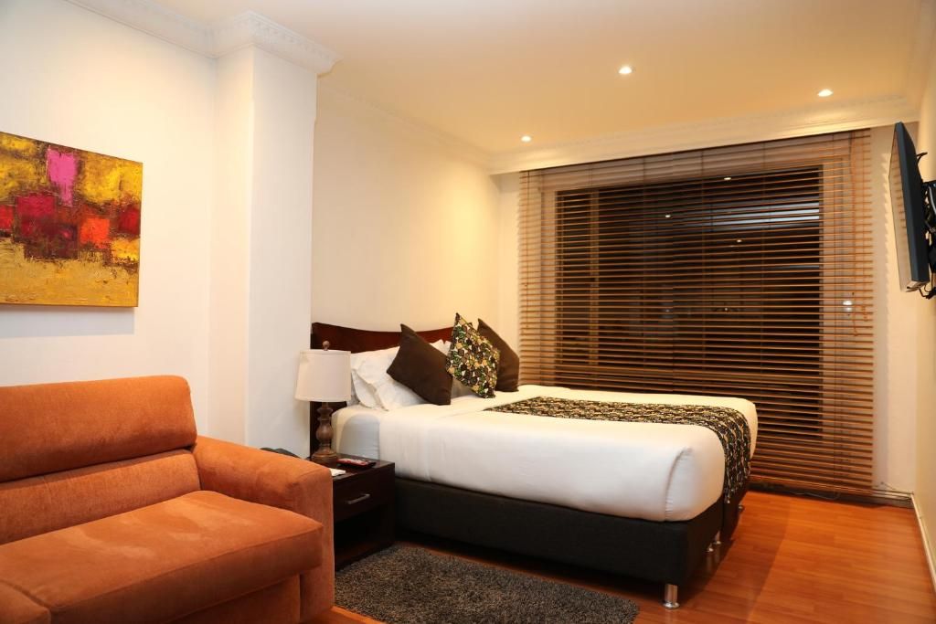 Une chambre d’hôtel avec un lit, une table de chevet, une lampe, un tableau au mur et une fenêtre avec des stores.