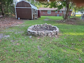 A circular stone fire pit in a grassy yard with a wooden shed and a brick house in the background. - The Hidden Gem — Cozy Southern Retreat (Hampstead)