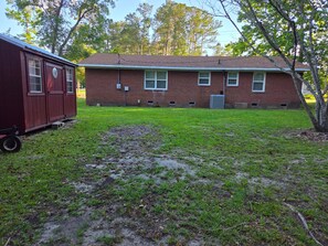 A single-story brick building with a flat roof, a small attached shed, and a grassy yard with a muddy patch. - The Hidden Gem — Cozy Southern Retreat (Hampstead)
