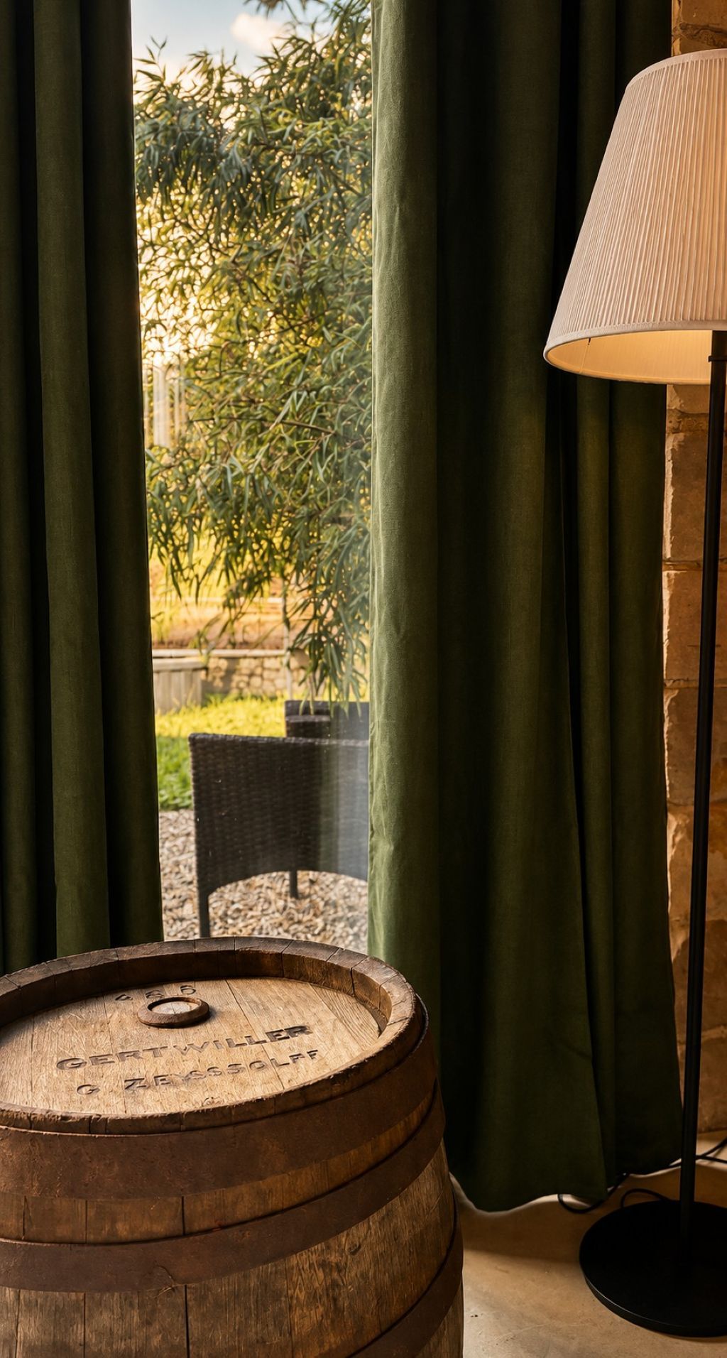 A wooden barrel, a lamp, and a view of greenery through curtains.