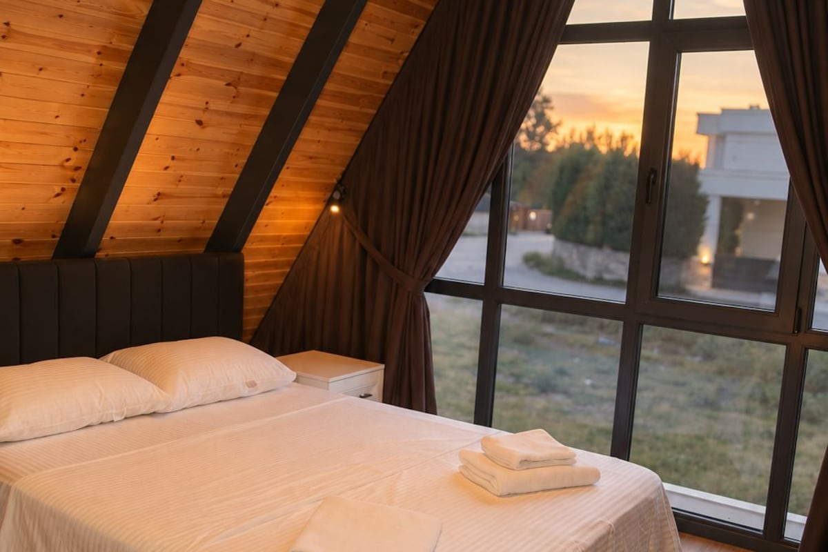 Un dormitorio con techo inclinado, una cama con ropa de cama blanca, una lámpara colgante y vistas al exterior a través de amplios ventanales.