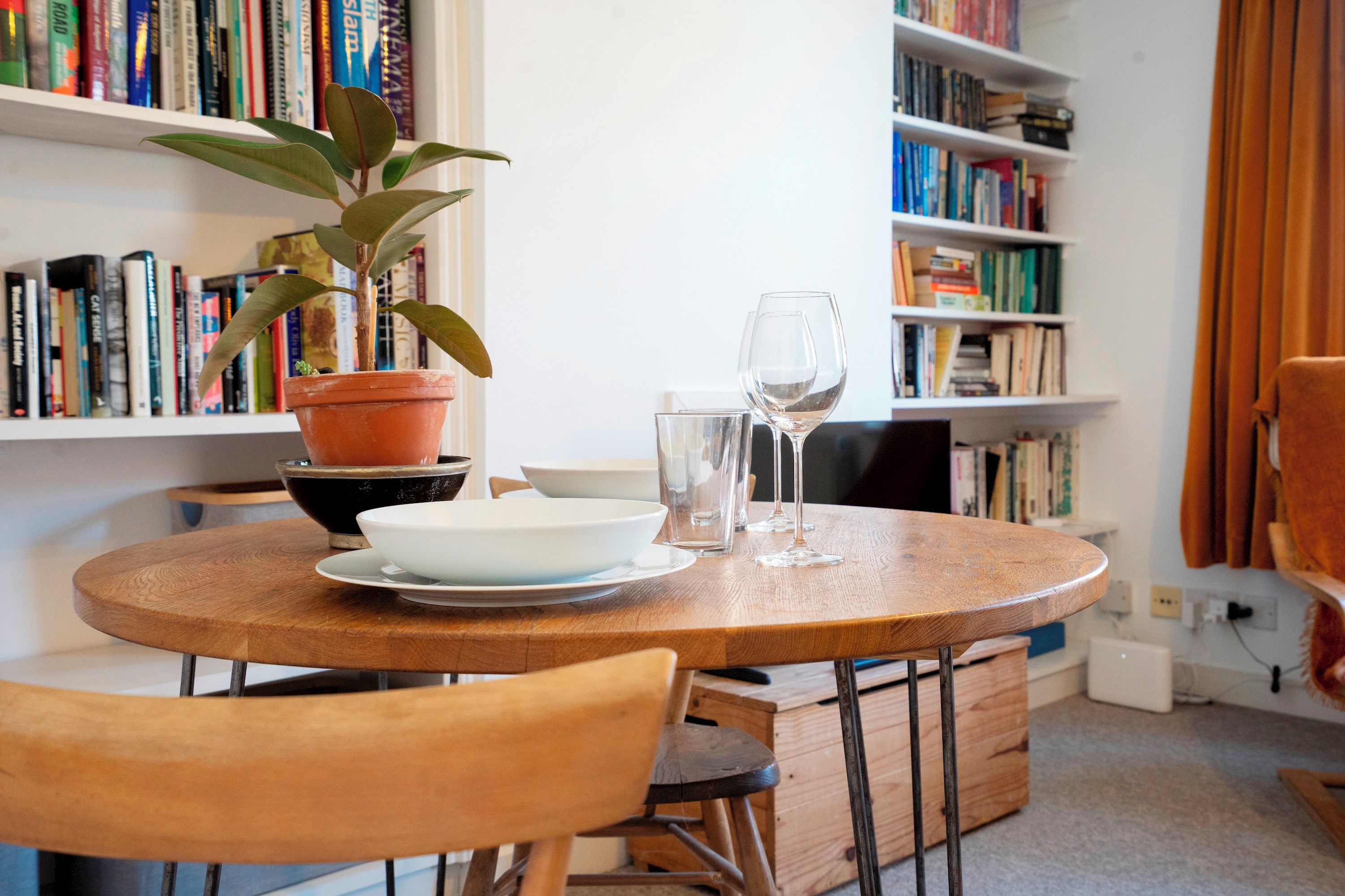 Une table ronde en bois, ornée d’une plante en pot, d’un bol blanc et de verres à vin.