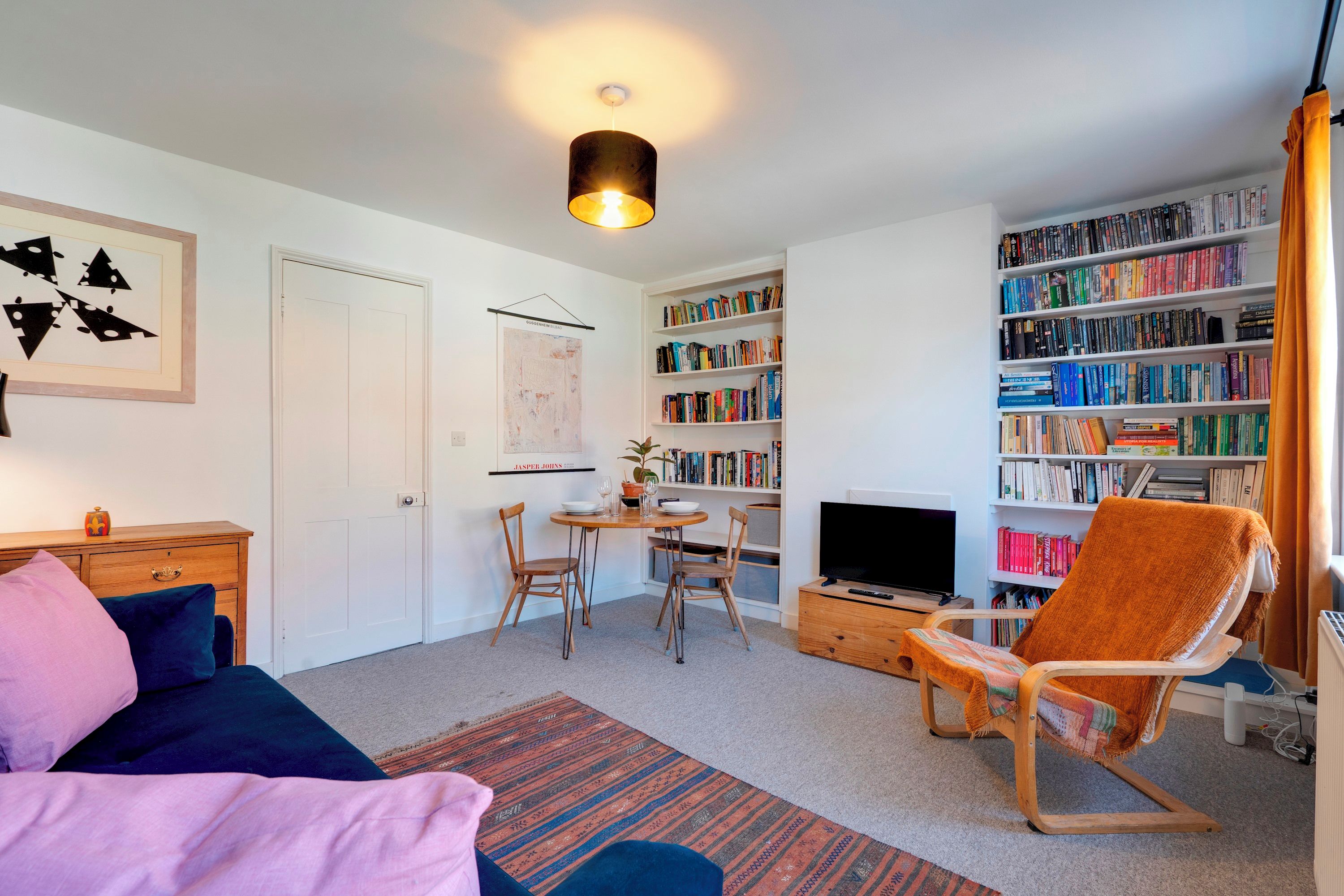 A well-lit living room with a sofa, a dining table with chairs, a bookshelf, a television, and a patterned rug.