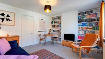 A well-lit living room with a sofa, a dining table with chairs, a bookshelf, a television, and a patterned rug.
