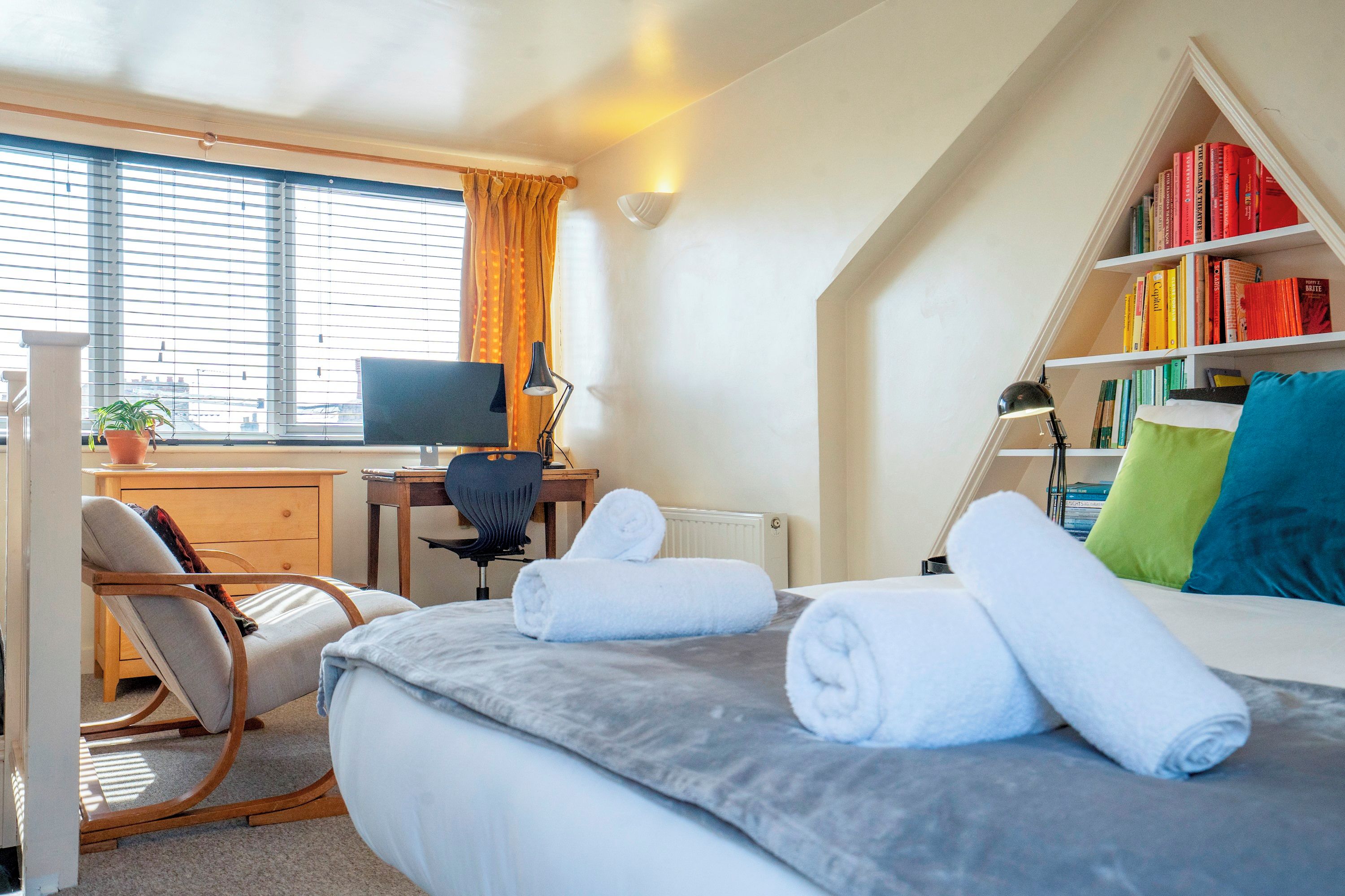 Une chambre à coucher avec un lit, un bureau avec un ordinateur, une chaise, une bibliothèque et une fenêtre avec des rideaux.