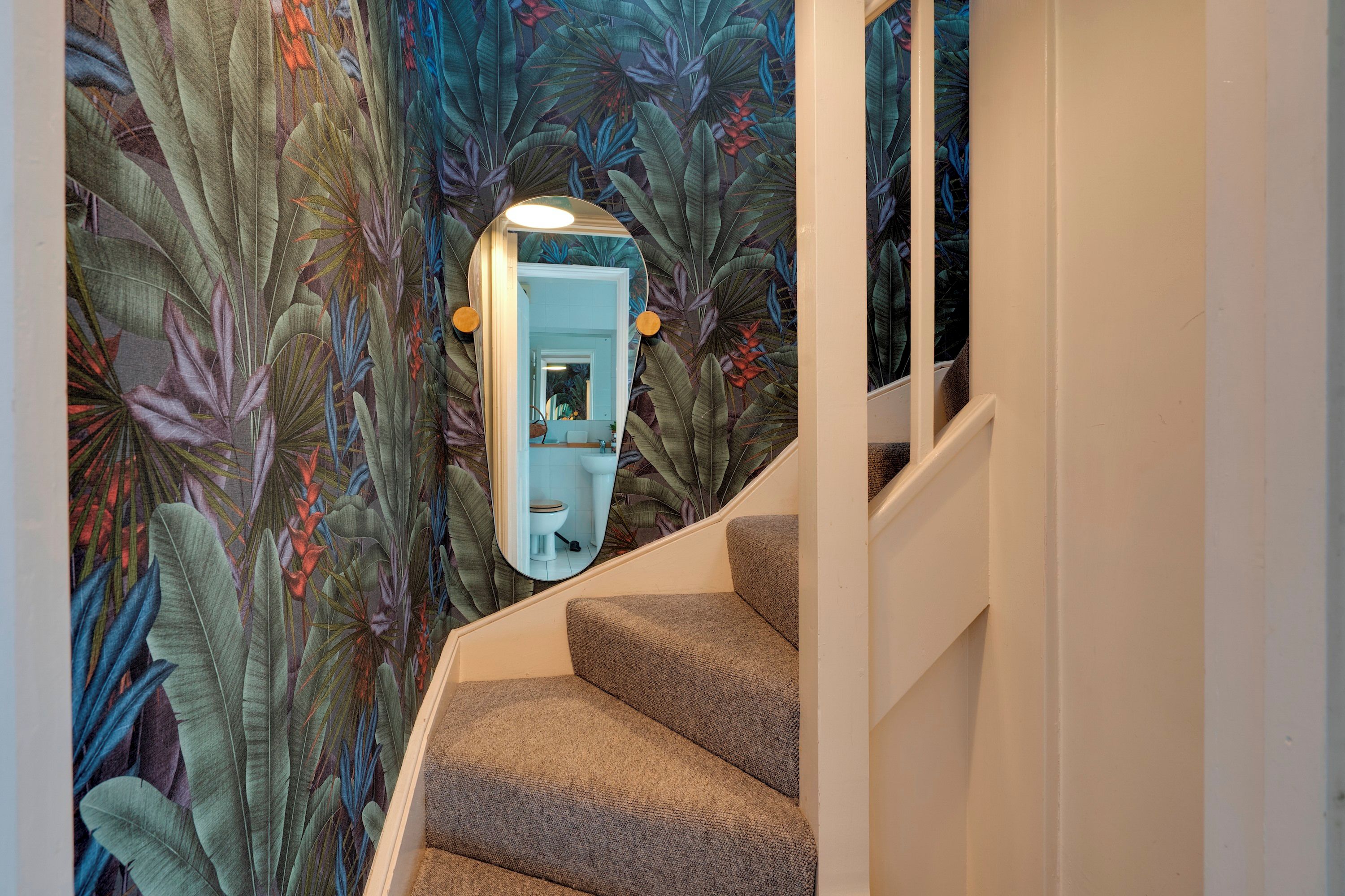 Un escalier avec un miroir qui reflète une salle de bain, le tout entouré d’un papier peint orné de motifs de feuilles tropicales.