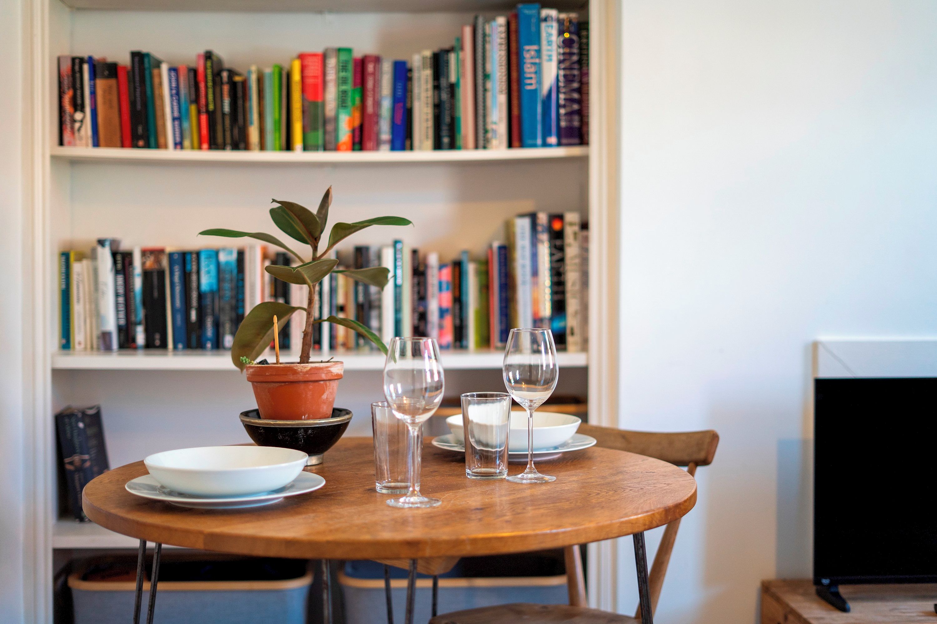 A round wooden table with four wine glasses and plates, a potted plant, and a bookshelf filled with books in the background.