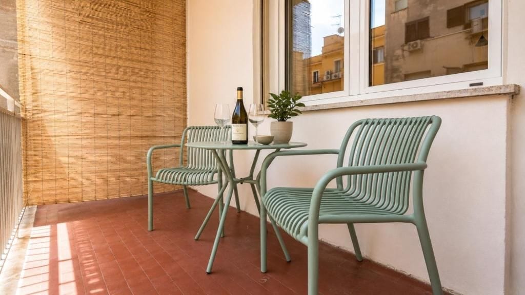 Un balcón con dos sillas de metal verde, una mesita, una botella de vino y una planta en maceta.