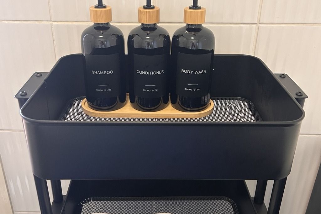 Una mensola in metallo nero con tre flaconi etichettati shampoo, balsamo e detergente corpo.