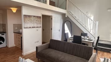 Een ruime woonkamer met een bank, een eettafel en een trap naar een hoger gelegen verdieping.