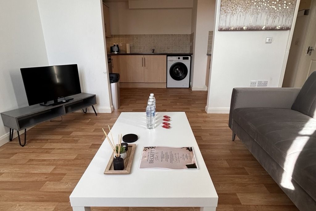 Una sala de estar con una mesa de centro blanca, un sofá gris, un televisor de pantalla plana y una zona de cocina al fondo.