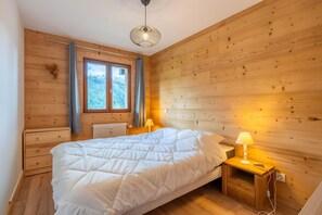 A wooden-paneled room with a bed, a window with a view of a forest, and a hanging light fixture. - Apartment 'Soladret' with Mountain View and Balcony (La Clusaz)