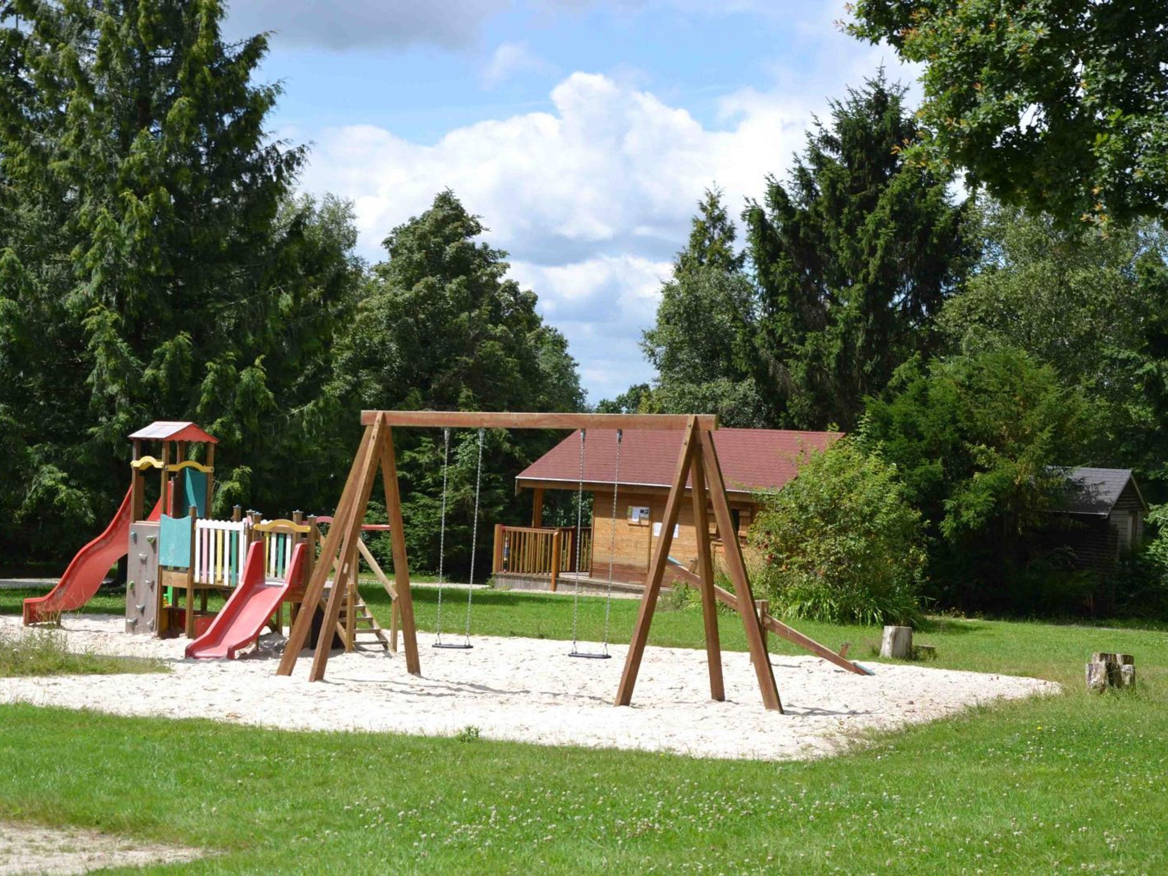 A playground with swings and slides, surrounded by trees and a building with a red roof.