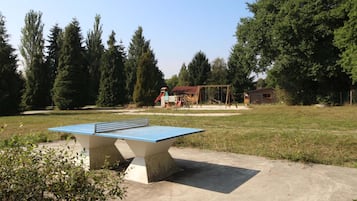 An outdoor ping pong table in a grassy area with trees and a swing set in the background.