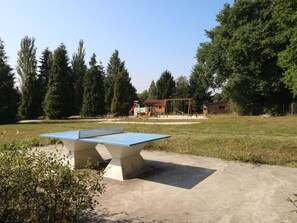 Una mesa de ping pong al aire libre en un césped con árboles y un columpio de fondo.