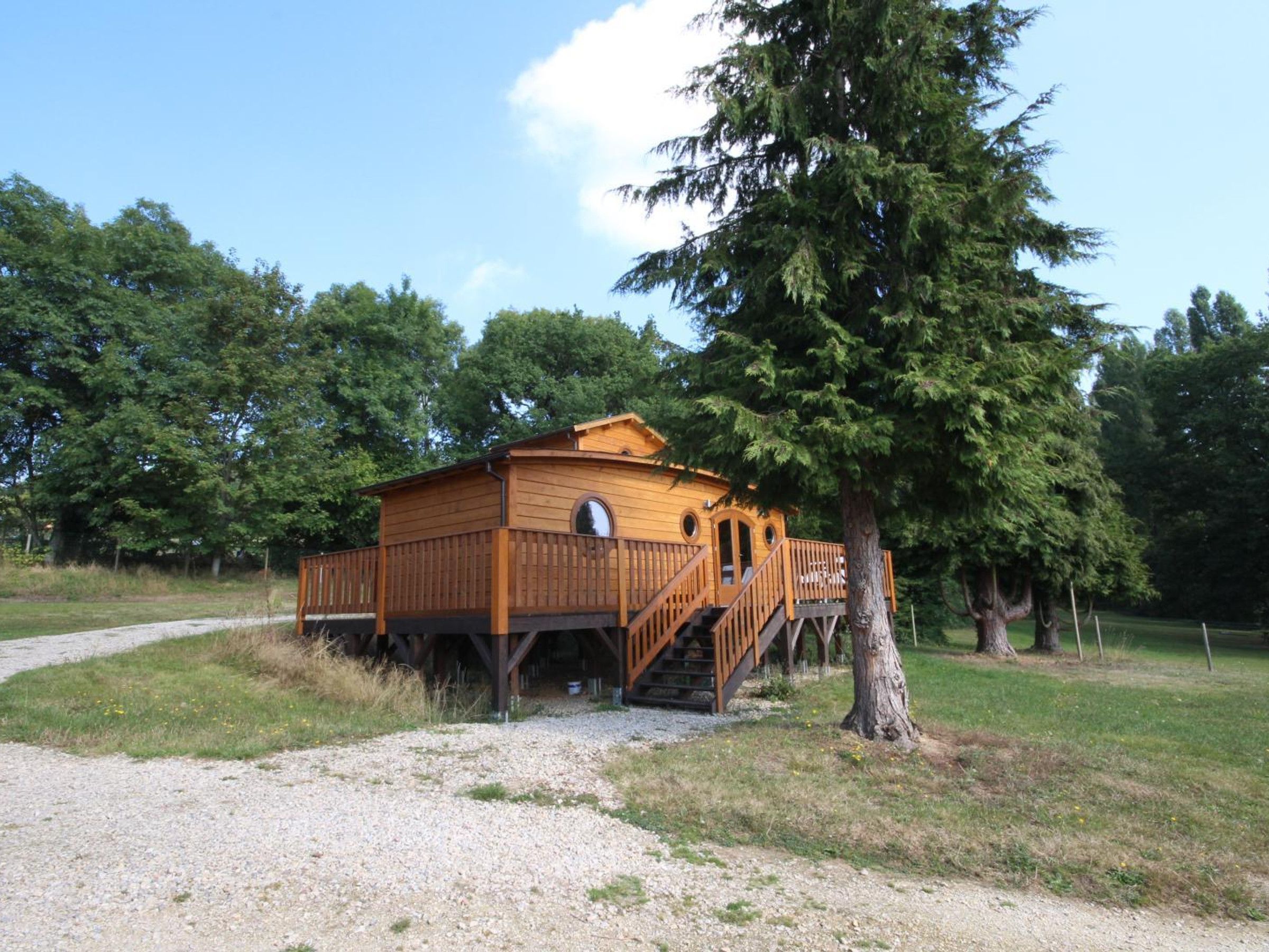 A wooden cabin with a deck and stairs, surrounded by trees and a gravel path.
