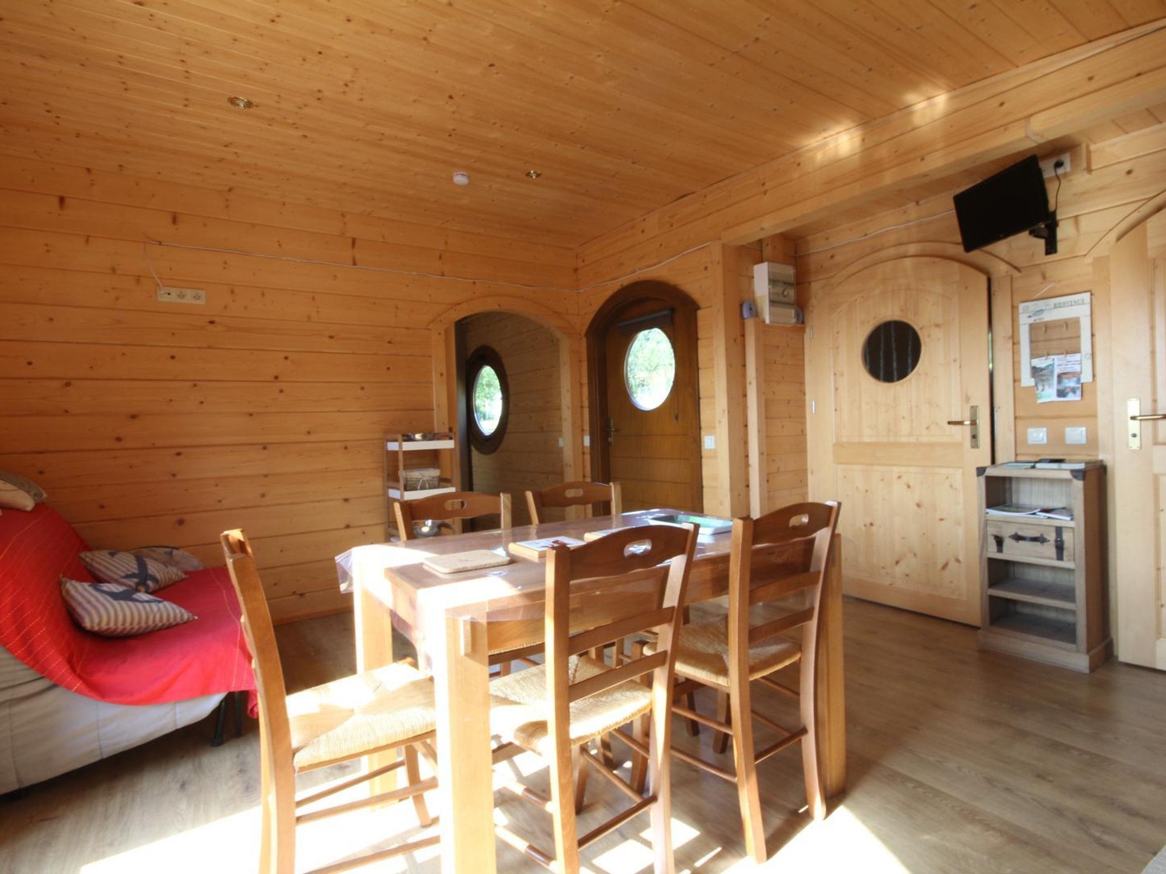 Interior d'una cabana de fusta amb una taula de menjador, cadires, un sofà amb una manta vermella i un armari de fusta amb una televisió.