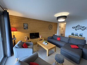 A modern living room with a sofa, armchair, and coffee table. A television is mounted on the wall, and there are decorative items on the shelves. - Apartment with south-facing balcony, mountain view - 6 guests (Auris)