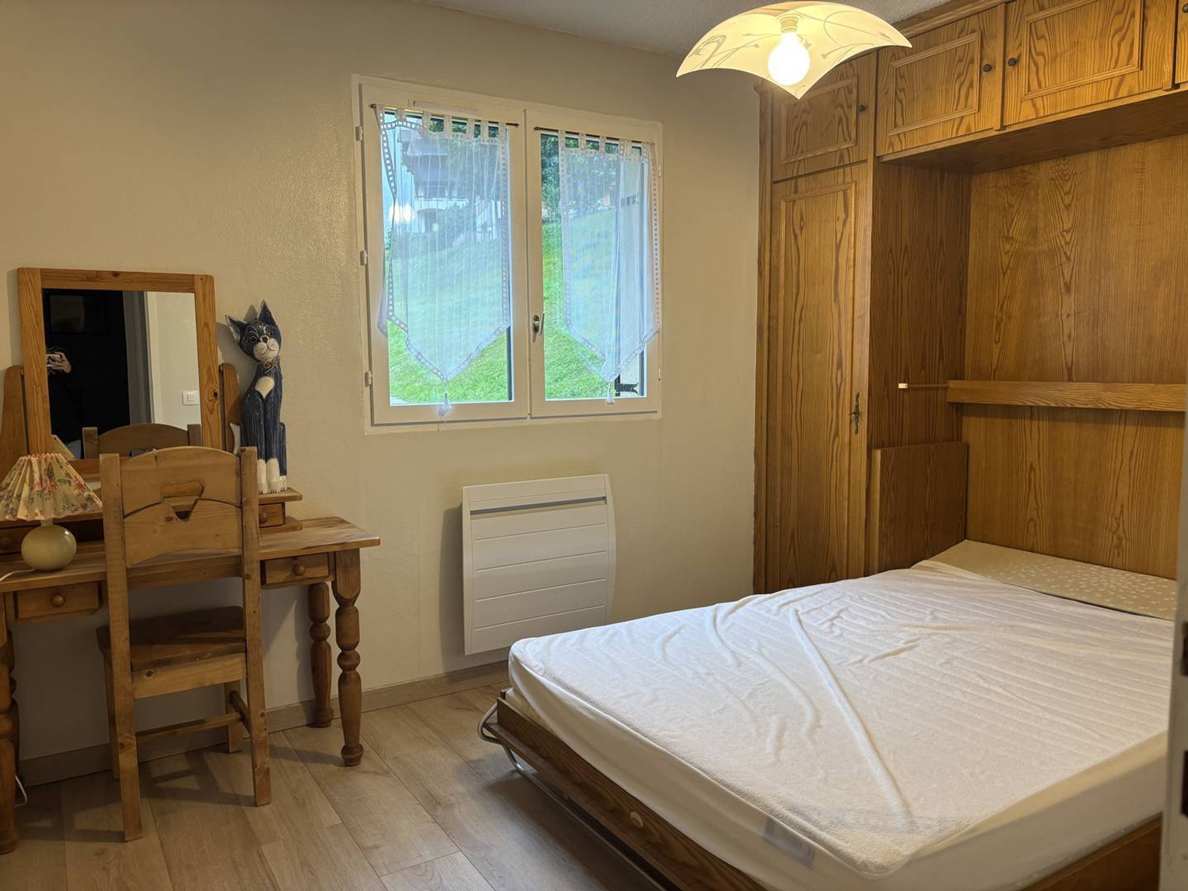 Een slaapkamer met een houten bureau, een stoel, een bed, een raam met gordijnen en een houten kledingkast.