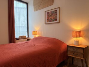 Ein Schlafzimmer mit einem Bett, zwei Nachttischlampen, einem Nachttisch, einer Schublade, einem Fenster mit Vorhängen und einem Wandbild.