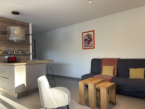 A modern living room with a dark sofa, a wooden coffee table, and a white chair. A kitchen area with a wooden countertop and a red toaster is visible. - Spacious apartment in the heart of Vercors - 6 guests (Villard-de-Lans)