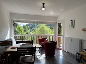 A modern room with a dining area, a red chair, and a view of a forest through large windows. - Cozy apartment mountain view - 4 guests (Villard-de-Lans)