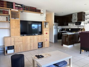 Ein modernes Wohnzimmer mit Flachbildfernseher, Holzsäcken, einem Essbereich mit Tisch und einer Küche mit dunklen Schränken und einer Mikrowelle.