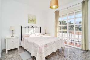 Une chambre à coucher avec un lit, des tables de chevet, une lampe, un tableau et une grande fenêtre avec des rideaux.