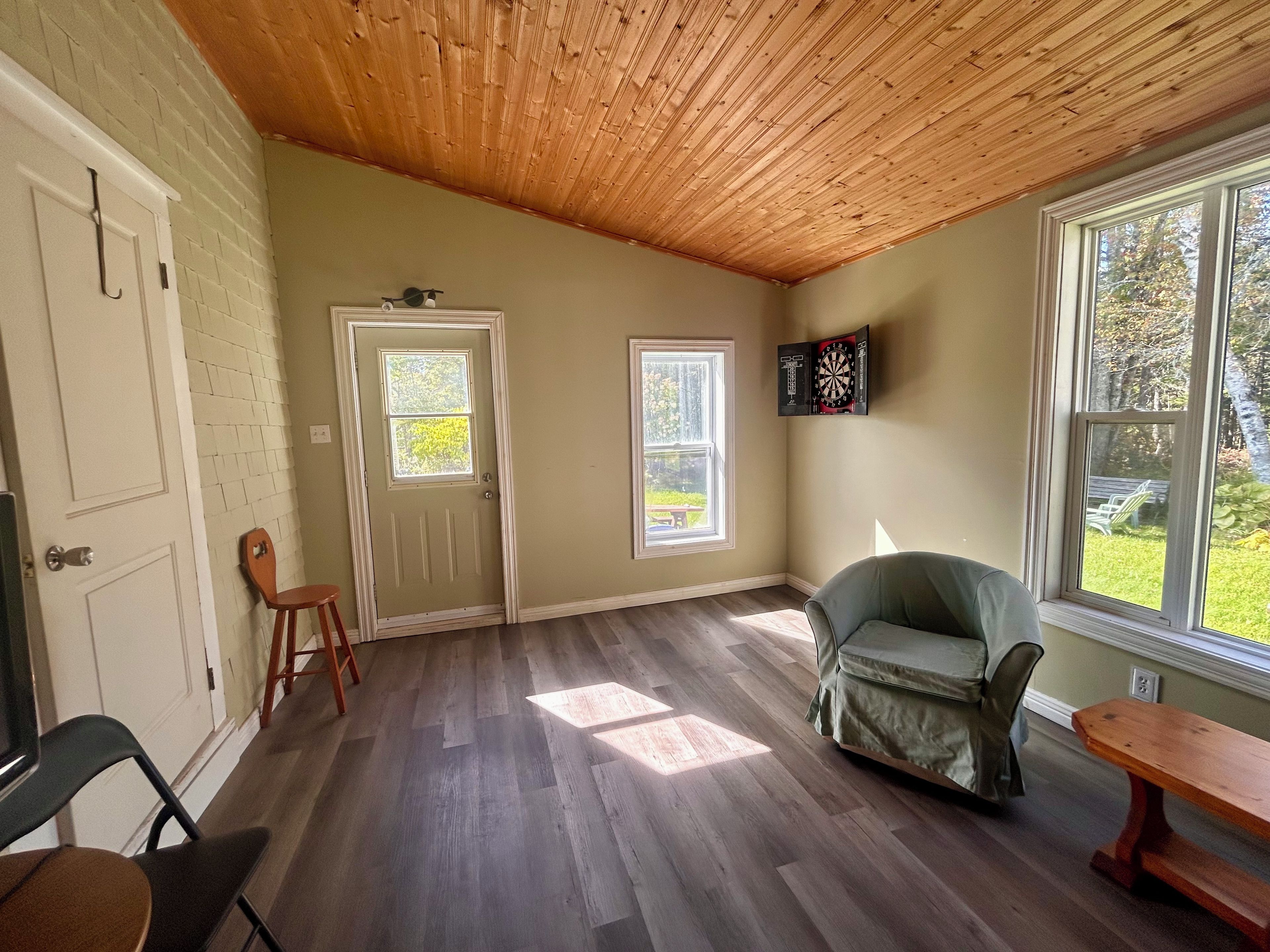 A room with a wooden ceiling, a door, a window, a dartboard, and a chair.