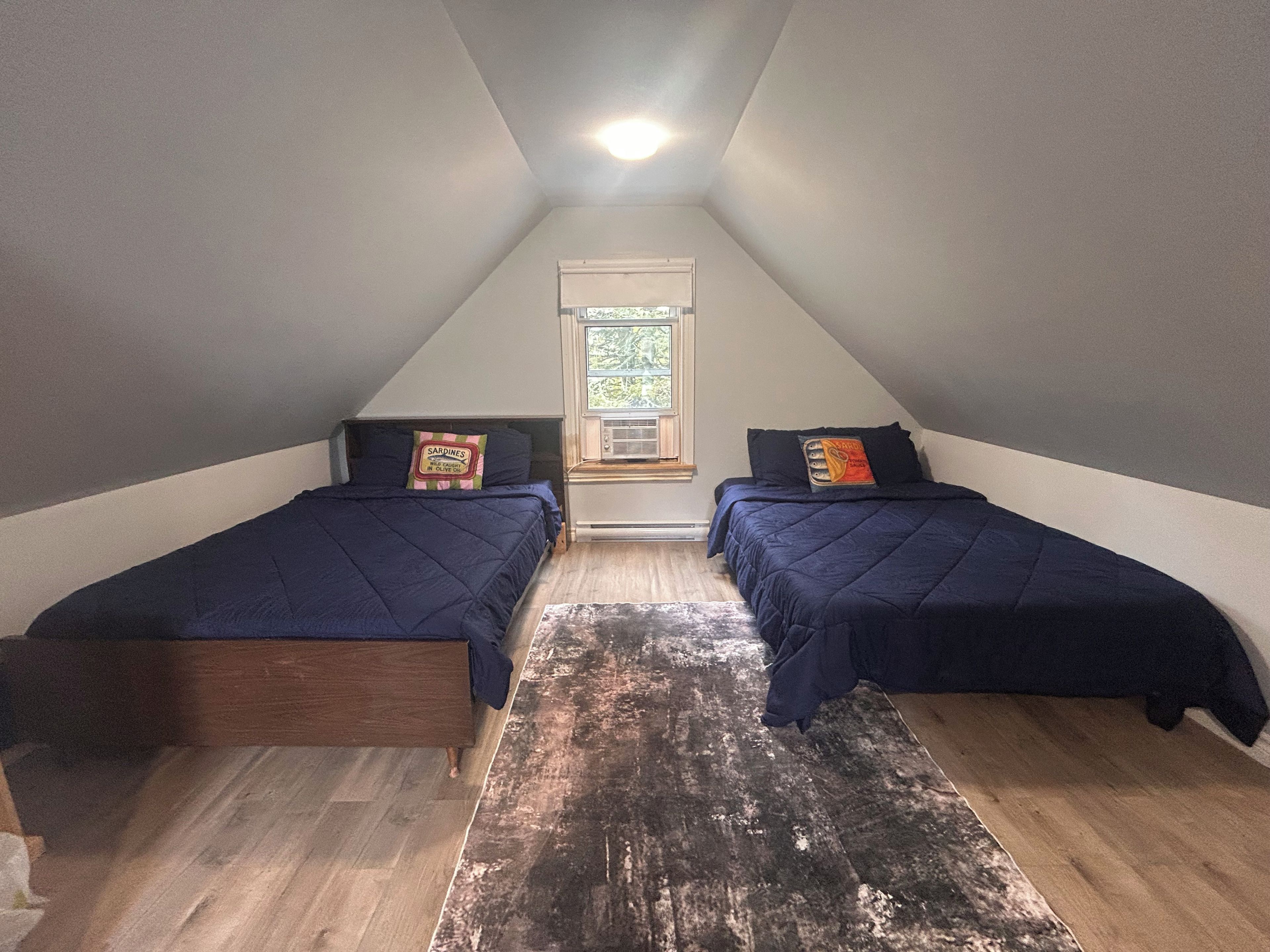 A small, triangular attic bedroom with two beds, a window with a view, and a ceiling light.