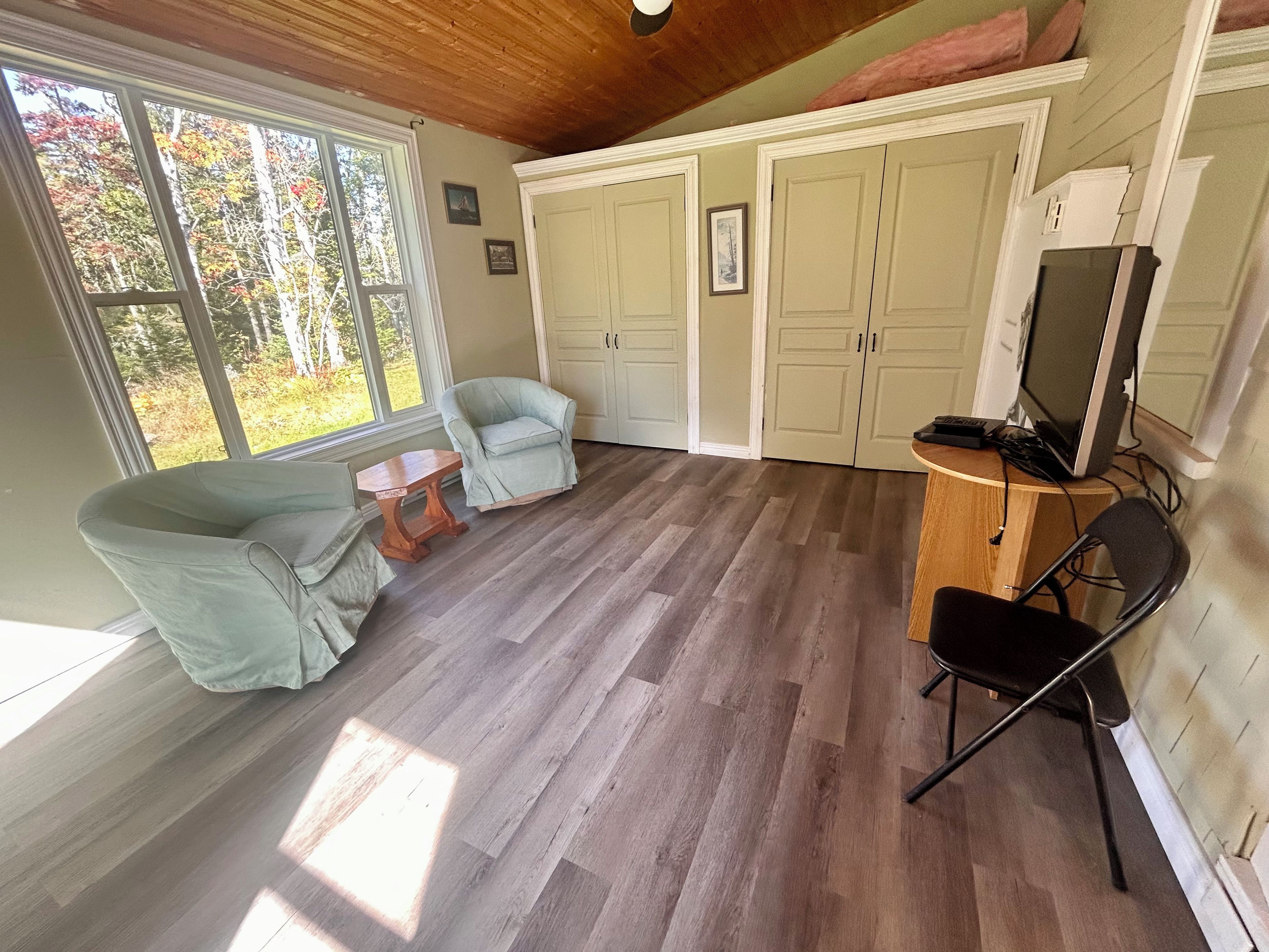 A room with a wooden ceiling, a large window with a view of trees, a light blue armchair, a wooden table, a TV on a stand, and a wardrobe.