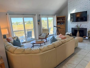 A living room with a large sectional sofa, a fireplace, a flat-screen TV, and a balcony with a view of the countryside. - New to our 2027 Inventory! Amazing views of Bass River Marina (West Dennis)