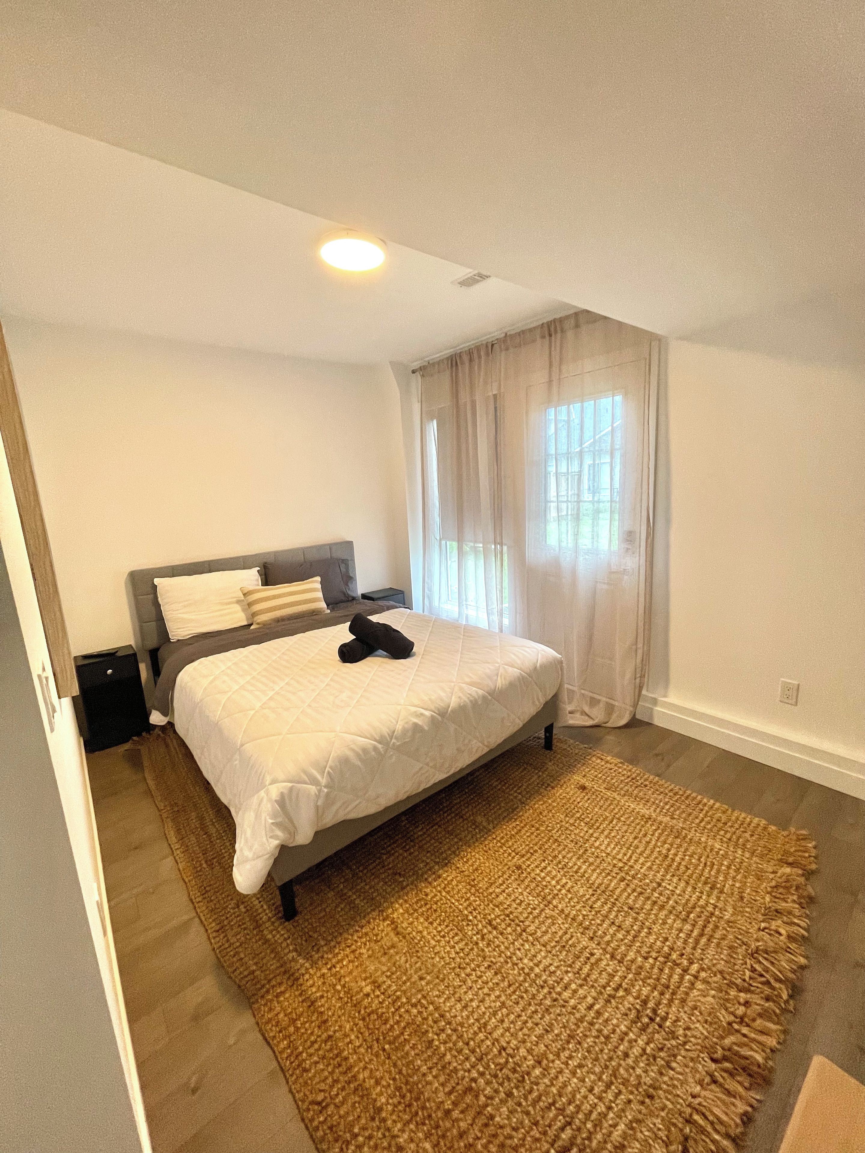 Une chambre à coucher comprenant un lit, une table de chevet, une commode, une fenêtre avec des rideaux et un luminaire au plafond.