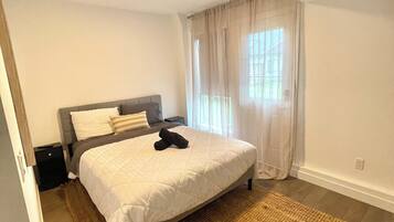 Une chambre à coucher comprenant un lit, une table de chevet, une commode, une fenêtre avec des rideaux et un luminaire au plafond.