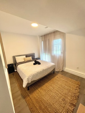 A bedroom with a bed, a bedside table, a nightstand, a window with curtains, and a ceiling light. - Canal Private Suite - Welland (Welland)