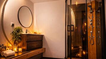 Een moderne badkamer met een douchecabine van glas, een houten wastafelmeubel, een ronde spiegel en kaarsen die sfeerverlichting creëren.