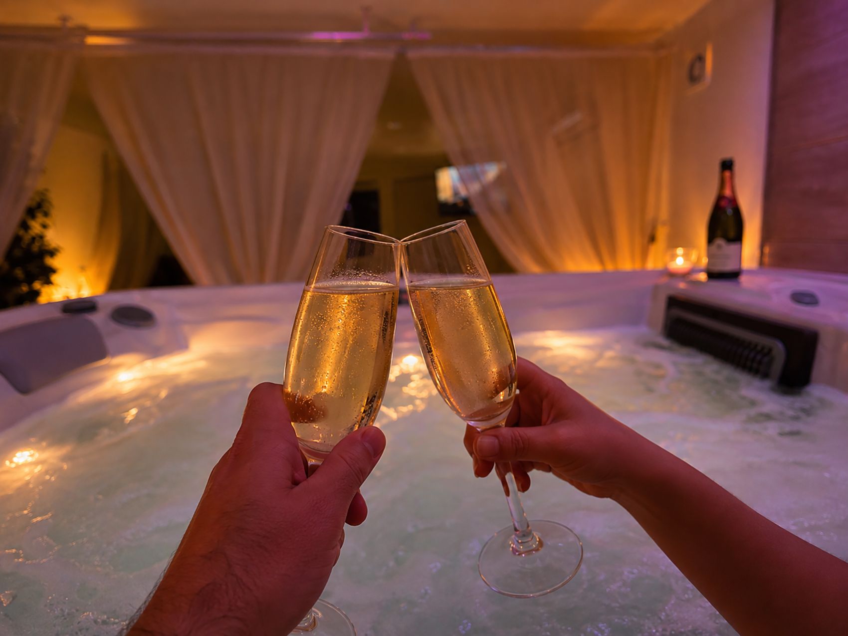Två händer som håller i champagneglas, ett bubbelbad och en vinflaska i bakgrunden.