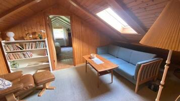 A cozy attic room with a wooden ceiling, a blue sofa, a wooden coffee table, a bookshelf, and a lamp.
