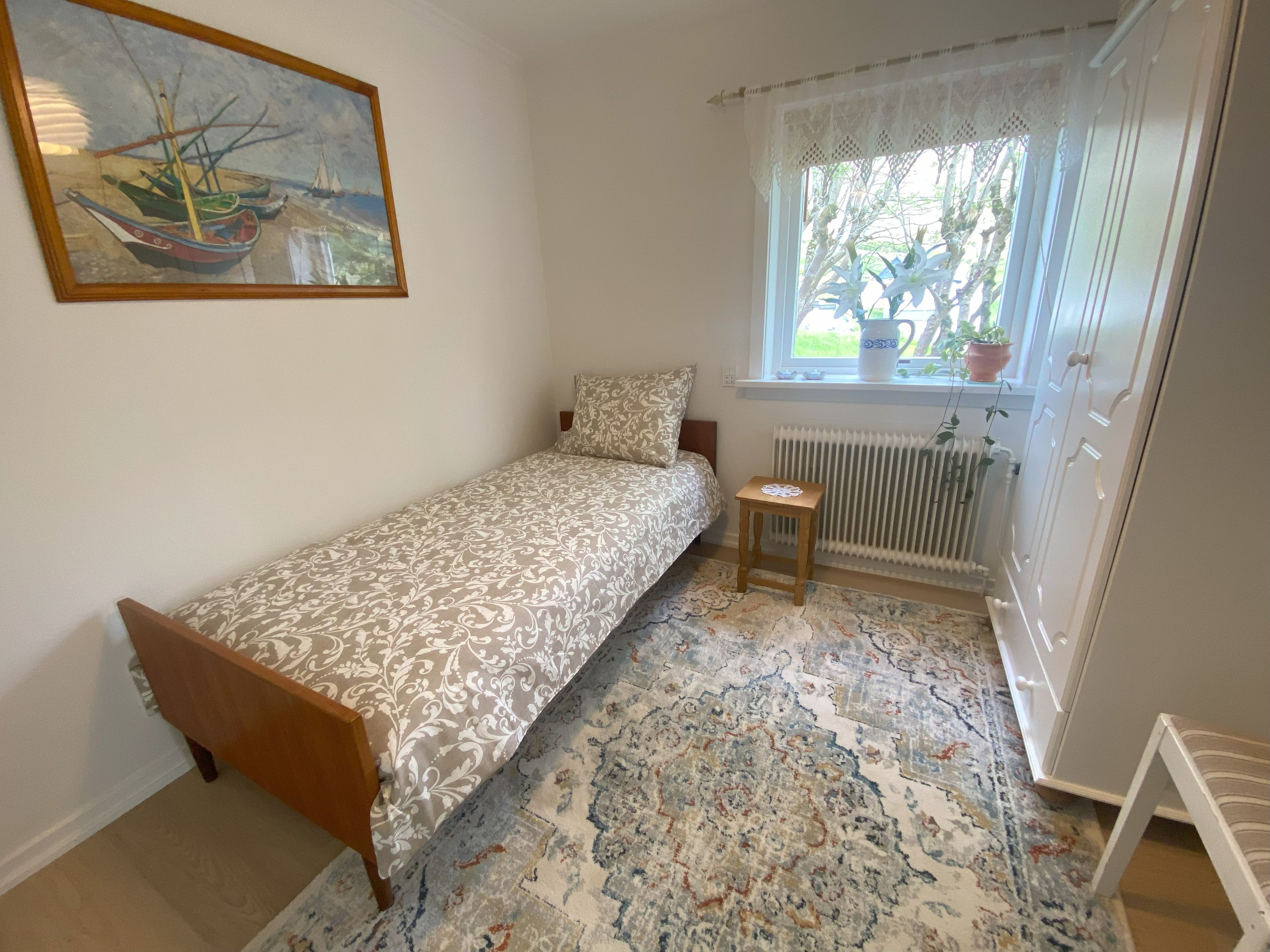 A small, single-bed room with a patterned bedspread, a small wooden stool, a radiator, and a window with a view of greenery.