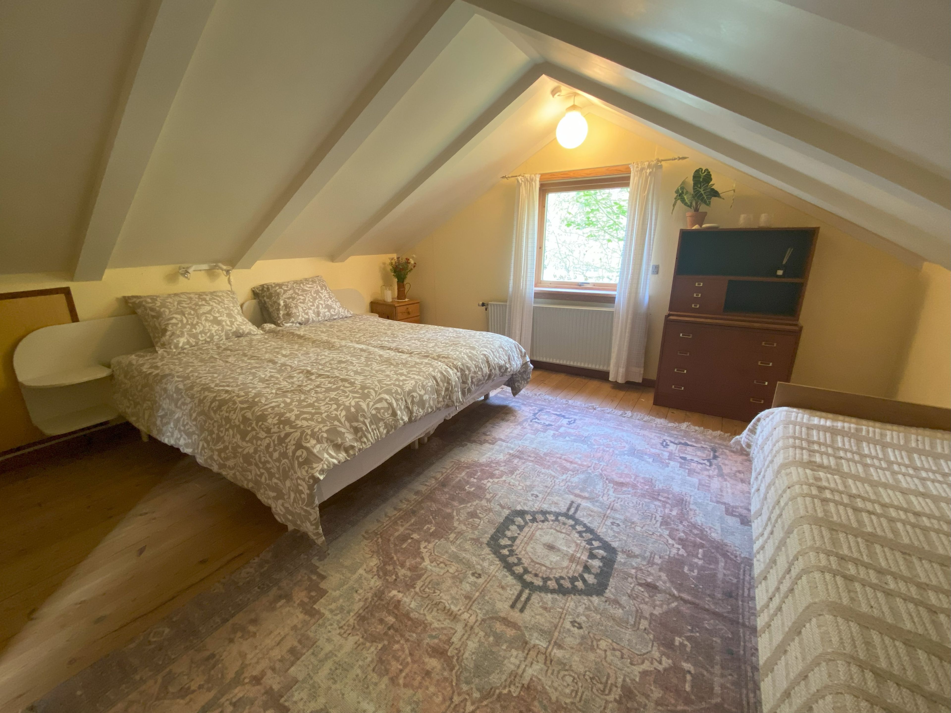 Quarto com cama, penteadeira, mesa de cabeceira, janela com cortinas e radiador.